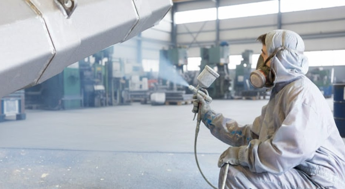 Person in protective gear spray painting a large industrial object in a workshop