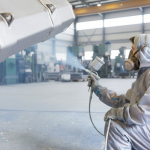 Person in protective gear spray painting a large industrial object in a workshop