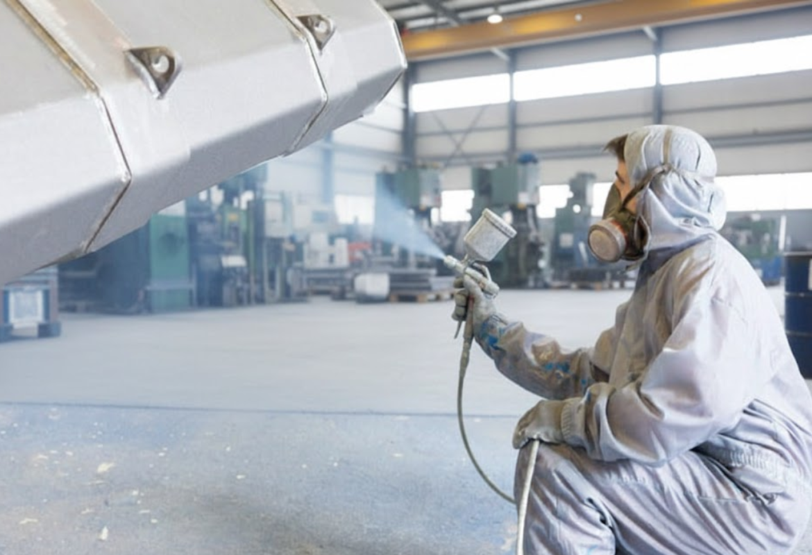Person in protective gear spray painting a large industrial object in a workshop